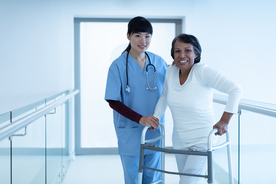 Medical assistant helping a patient walk with a walker in a hospital setting.