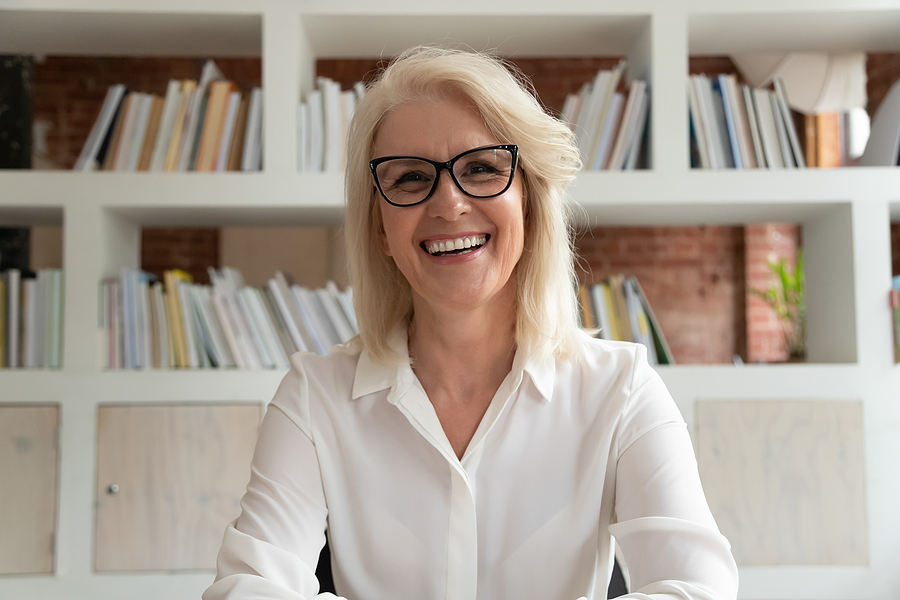 Mature teacher smiling at her desk in front of a bookshelf.