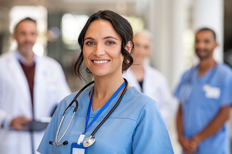 Smiling female nurse standing in front of a medical team, background is blurry but you can tell they are medical professionals.