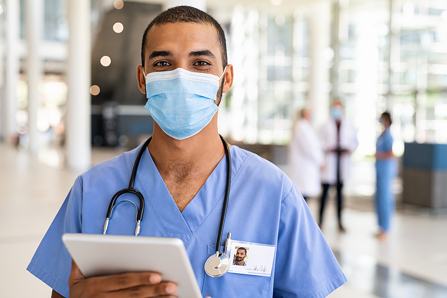 Medical assistant wearing a surgical mask smiling at the camera while holding an electronic tablet.