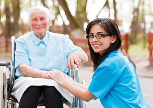 Nurse wearing blue scrubs helping an elderly lady in a wheelchair. 