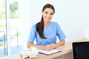 Young medical assistant wearing blue scrubs is writing on a clipboard in a medical office. 