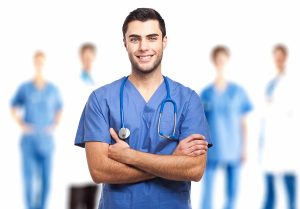 Male medical assistant wearing blue scrubs posing with his arms folded with medical workers blurred in the background. 