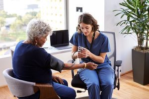 Female medical assistant wearing dark blue scrubs taking a senior female patient's blood pressure in an office setting. 