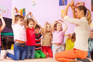 Smiling female preschool teacher sitting in front of a large group of young students leading an activity. 