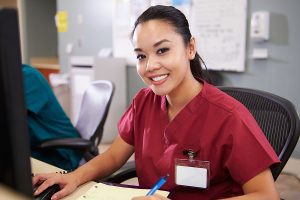 Female medical assistant wearing burgundy scrubs and smiling in front of a computer. 