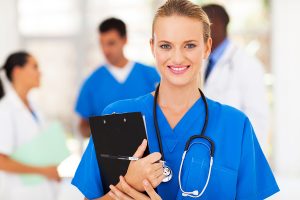 Female healthcare worker wearing blue scrubs holding a clipboard and smiling at camera with blurred out healthcare workers in the background. 