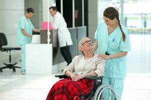 Female nurse wearing light green scrubs helping an older female patient in a wheel chair. 