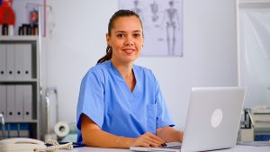 Female medical assistant wearing blue scrubs sitting in front of a laptop offering telemedicine support. 