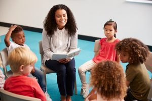 Female teacher reading to a group of young students, seated in chairs in a circle. 