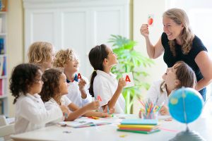 Smiling female teacher holding up a flashcard to a group of young students wearing white school uniforms. 