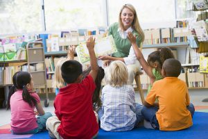 Female teacher reading a story to a group of children sitting on the floor in front of her. 