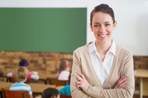 Female teacher standing in the back of her classroom, smiling at the camera. 