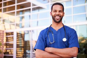 Male healthcare worker wearing blue scrubs and smiling at the camera. 