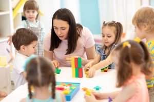 Female teacher sitting at a table with young students, doing an activity with blocks. 