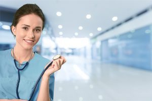 Female medical assistant wearing teal scrubs and holding a stethoscope in a white hallway. 