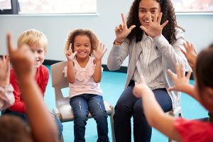 Female teacher with a group of preschool aged children sitting on chairs in a circle, and holding up their fingers doing a math exercise. 