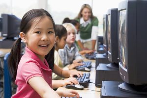 Young child smiles at the camera while typing on a computer with other classmates with teacher in the background. 