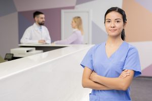 Female medical assistant wearing blue scrubs standing in front of a desk with doctor and patient in the background. 