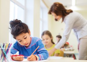 Young female student holding a pencil and working on an assignment at her desk with female teacher blurred out behind her. 