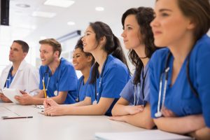 Group of medical students wearing blue scrubs and sitting at a table listening to a classroom lesson. 